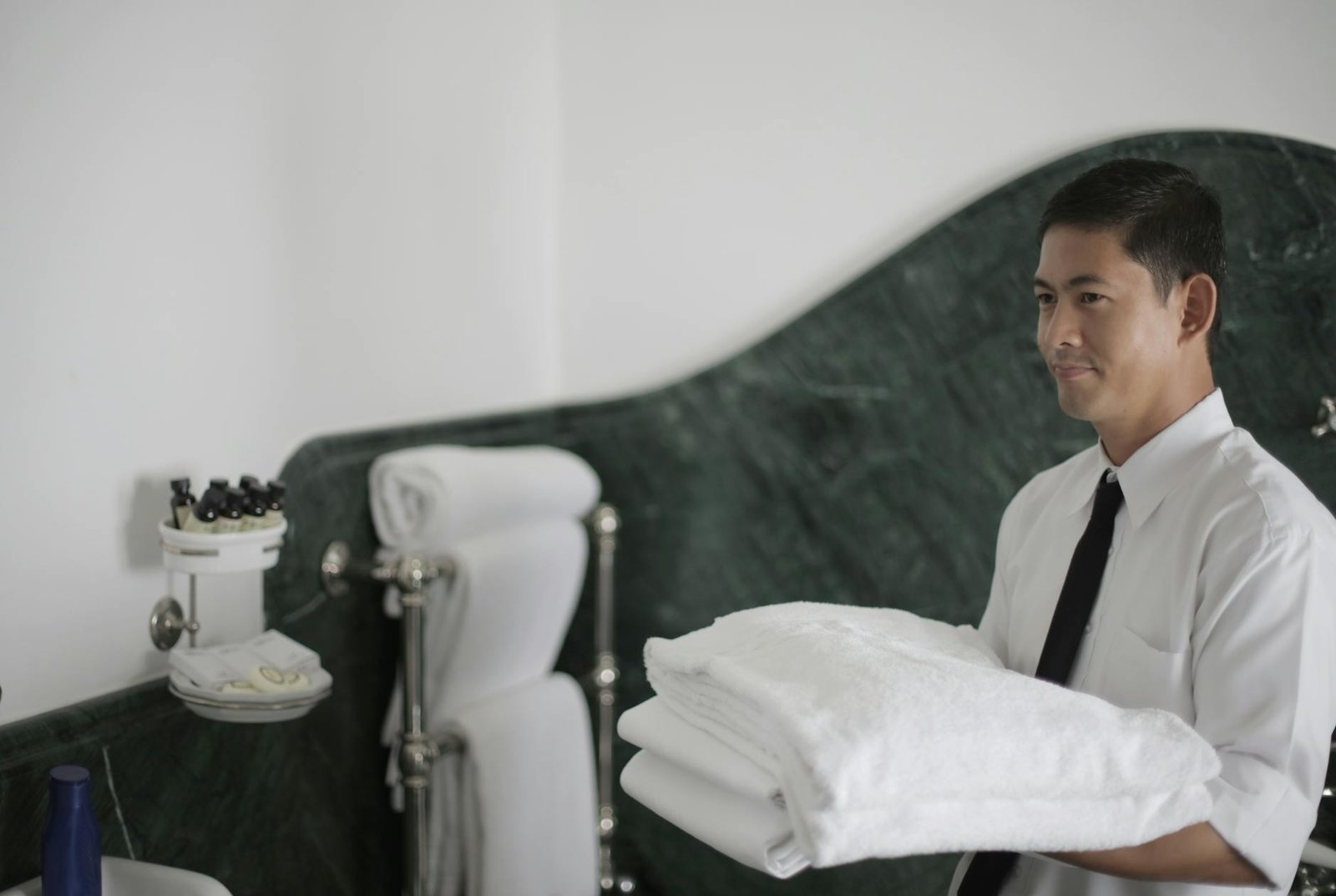 Side view of smiling ethnic male hotel worker holding stack of clean white towels while cleaning bathroom